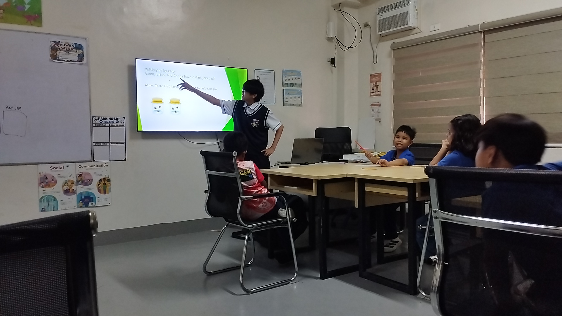 A PCIS student explaining a multiplication problem to younger learners during the Teacher-for-a-Day activity while classmates listen and participate, illustrating the preparation, communication, and engagement involved in teaching.